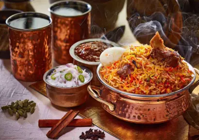Aromatic mutton biriyani served with eggs, gravy, raita garnished with spices, and glasses of water.